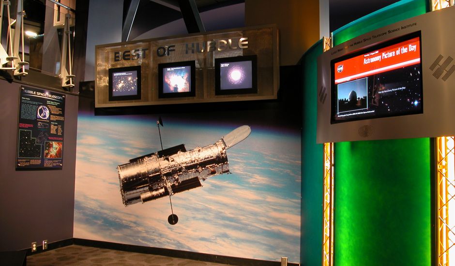 Museum exhibit with large image of Hubble over Earth below sign reading Best of Hubble with three monitors showing different images. Another poster and monitor display are visible.