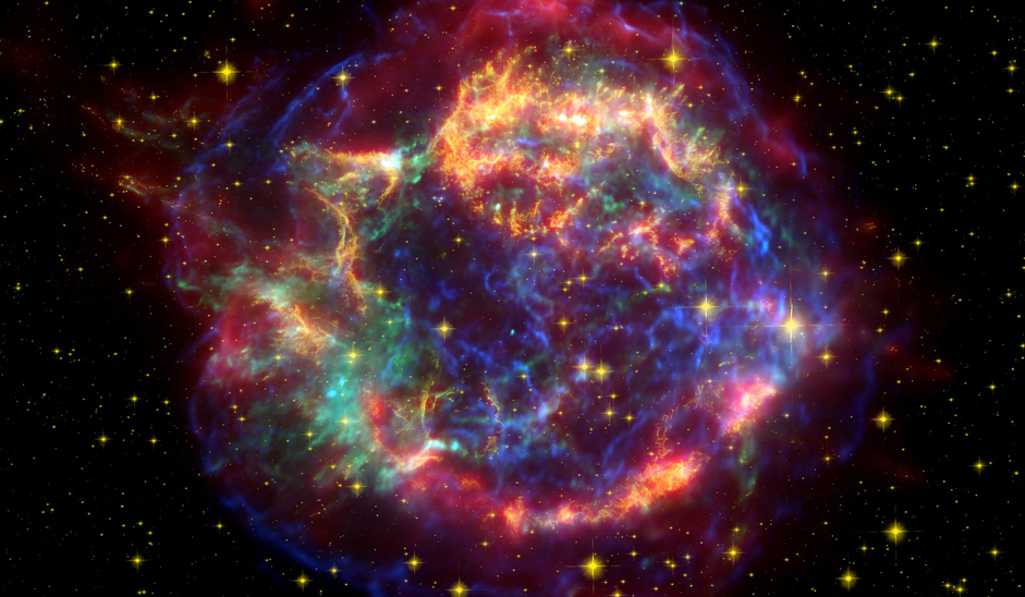 Large, colorful, semi-transparent circular object on a background of space scattered with small orange points of light. The large object has a complex cloudy and filamentous texture, with regions of blue, green, orange-yellow, and red-purple.