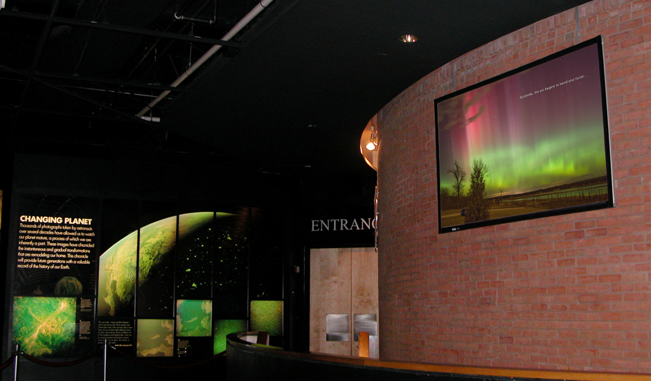 Photograph of a ViewSpace video playing on a large monitor mounted on a brick wall outside of an exhibit hall.