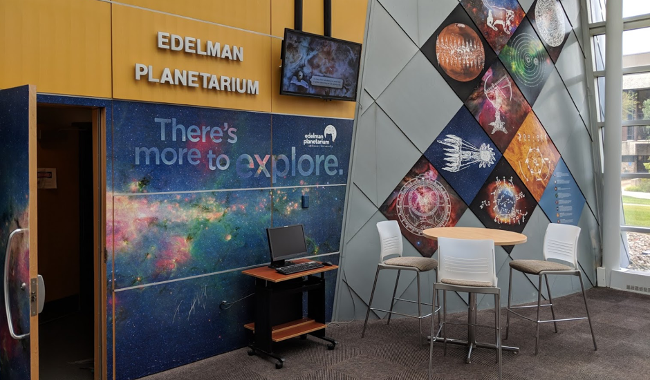 Lobby and open door of dark room labeled Edelman Planetarium with images of space covering wall panels, that also read There's more to explore. Lobby includes small table and chairs and cart with computer station.