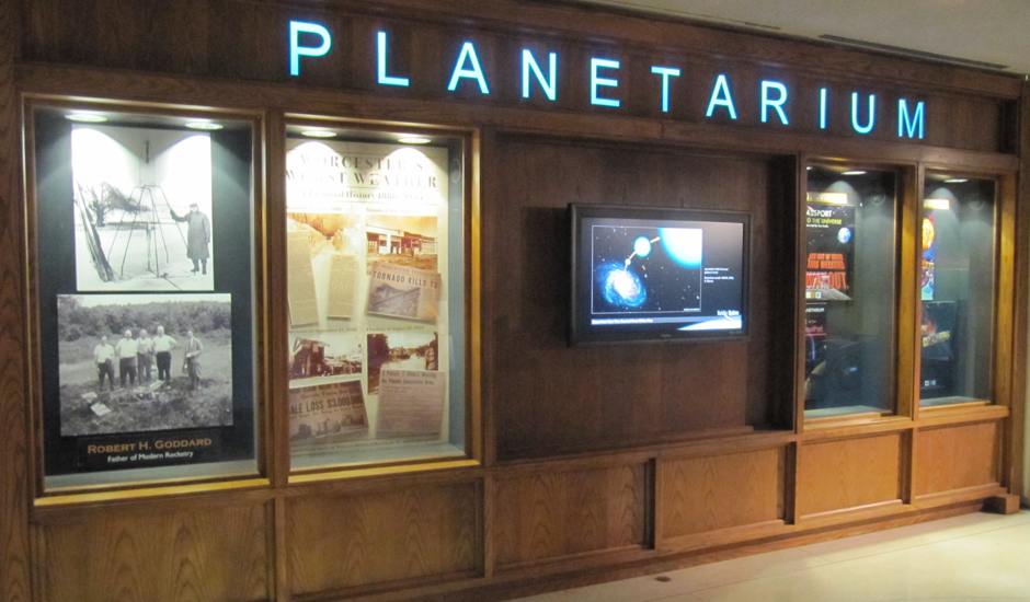 Photograph showing a monitor with a ViewSpace astronomy video playing, mounted at eye-level on a wall beneath the word, "Planetarium." The monitor sits between two sets of glass-fronted cases displaying historical photos and newspapers, and images and posters about space.
