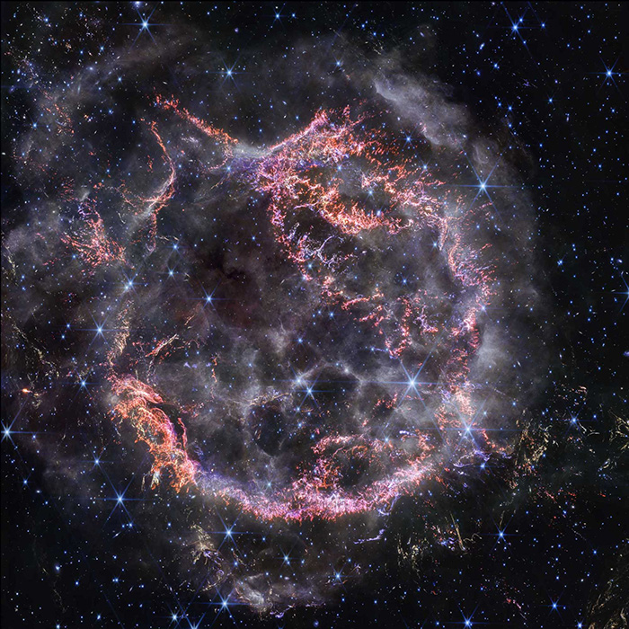 Cassiopeia A, a circular-shaped cloud of gas and dust with complex structure. The inner shell is made of bright pink and orange filaments studded with clumps and knots that look like tiny pieces of shattered glass. Around the exterior of the inner shell, particularly at the upper right, there are curtains of wispy gas that look like campfire smoke. The white smoke-like material also appears to fill the cavity of the inner shell, featuring structures shaped like large bubbles. Around and within the nebula, there are various stars seen as points of blue and white light.