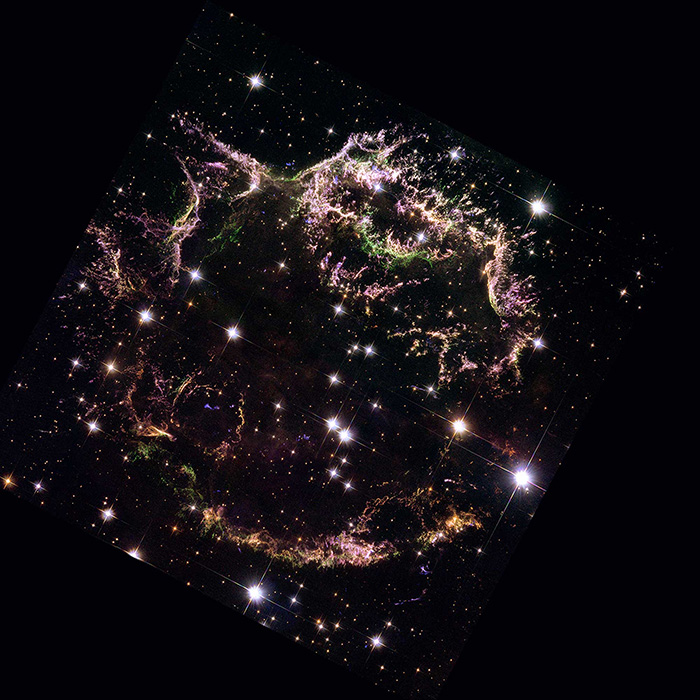 The Cassiopeia A supernova remnant with bright and dim stars scattered across the black background. The nebula is roughly circular, made of sparse green and purple thread-like filaments with pink edges. There are no filaments in the center of the remnant, allowing the starry background to shine through.