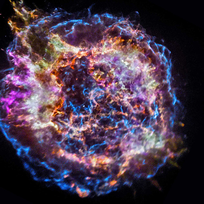Cassiopeia A supernova remnant, showing a roughly circular cloud of multicolored gas against a black background. The colors range from blue and purple to pink and yellow, resembling a cosmic smoke ring. The outside of the remnant appears as a mottled blue ring. Inside of the blue ring is another wide ring, made of a series of dense filaments in shades of pink, white, and orange. The center of the remnant is dominated by a network of blue filaments. There is a bright white point near the center.