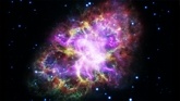 Image of the Crab Nebula showing large loops of purple, yellow, and green that form a misshapen oval set against the black background of space.