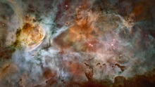 Image of the Carina Nebula: wispy colors of gas and dust appear in a range of gray-blues and rusty reds with a few pink stars dotting the scene.